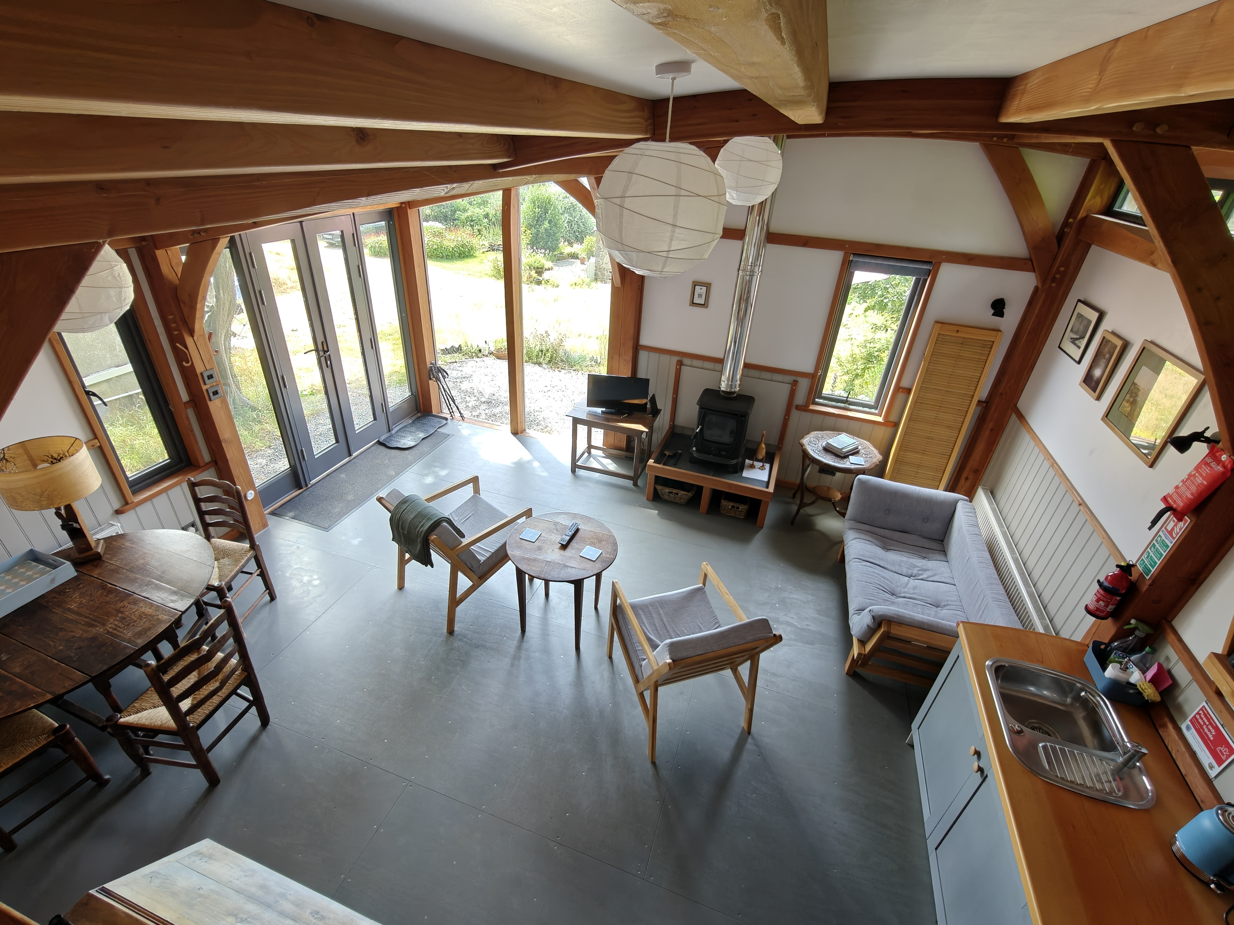 A view from the height of the living area of a wooden cabin, showing chairs, wood burner, TV, sofa bed, kitchenette and dining table.